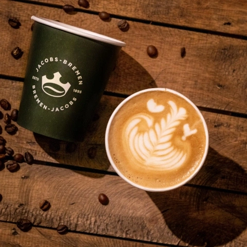 Kaffeebecher mit Latte Art und Jacobs-Bremen-Logo auf Holzuntergrund