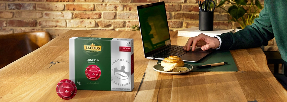 Kaffee-Kapseln, Laptop, Gebäck und eine Person, die an einem Holztisch arbeitet.