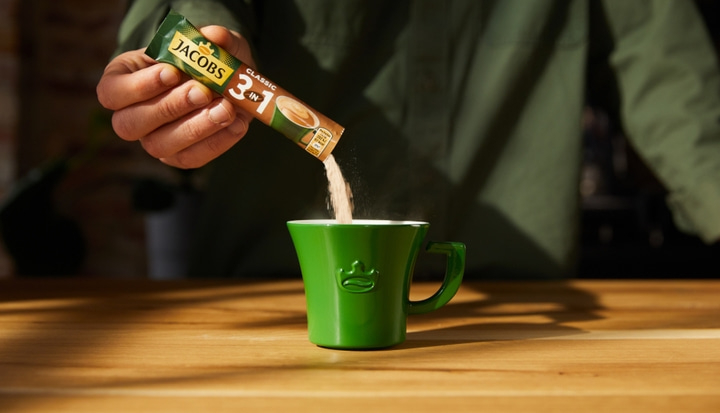 Person gießt Jacobs 3-in-1-Kaffee in eine grüne Tasse auf einem Holztisch.