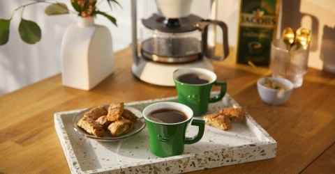 Zwei grüne Kaffeetassen mit Kaffee auf einem Tablett mit Gebäck und Kaffeemaschine.