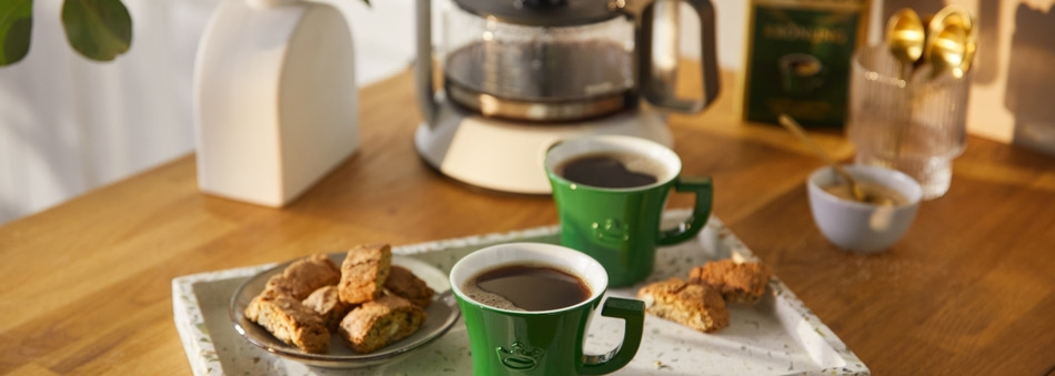 Zwei grüne Kaffeetassen mit Gebäck auf einem Tablett, Kaffeemaschine im Hintergrund.