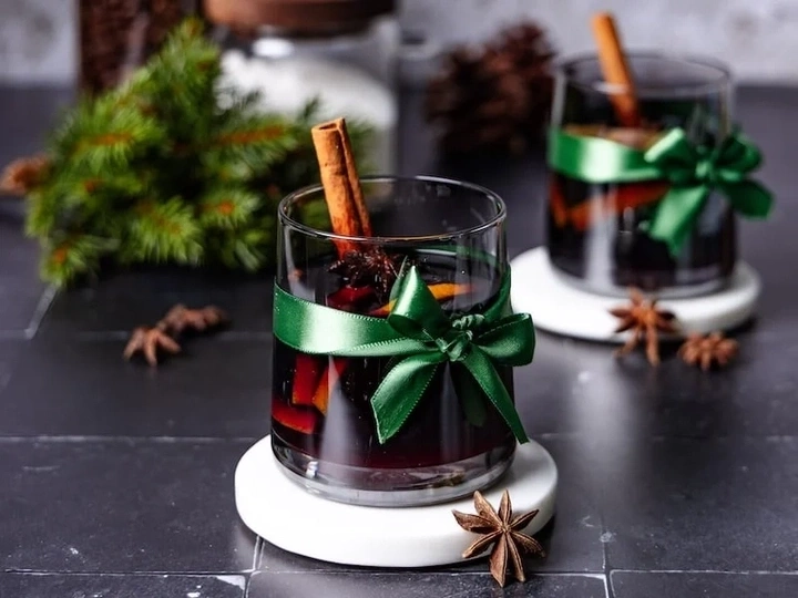 Zwei Gläser Glühwein mit Zimtstangen und Sternanis, dekoriert mit grünen Schleifen.